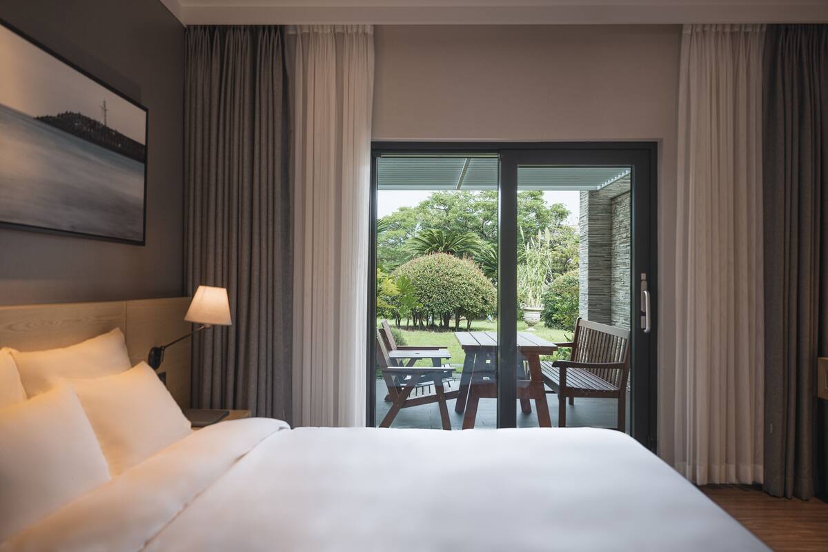 A hotel room with a large bed, a nightstand, a lamp, and a view of an outdoor patio with a table and chairs.
