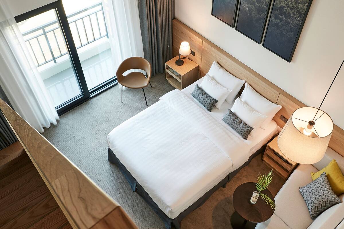 A hotel room with a large bed, a bedside table with a lamp, a chair, a small table with a plant, and a view of the outside balcony.