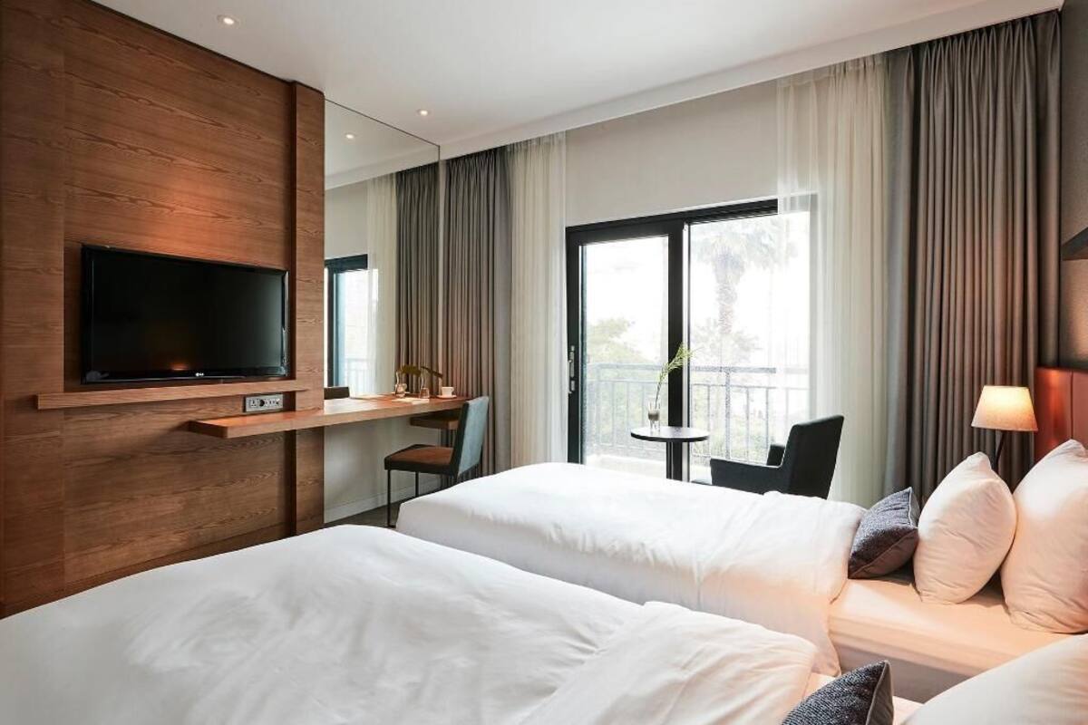 Habitación de hotel con dos camas, un televisor montado en la pared, un escritorio con una silla y un balcón con cortinas.