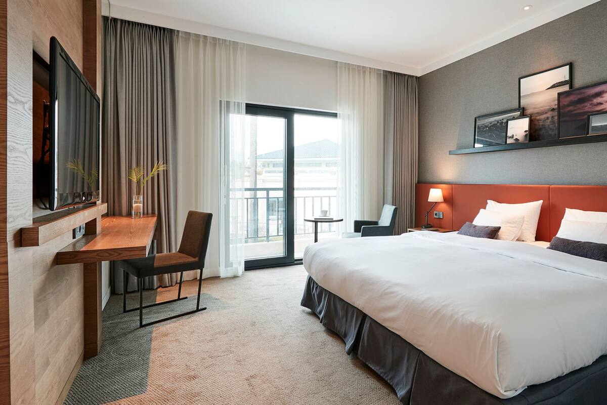 A hotel room with a large bed, a desk with a chair, a TV, and a balcony with curtains.
