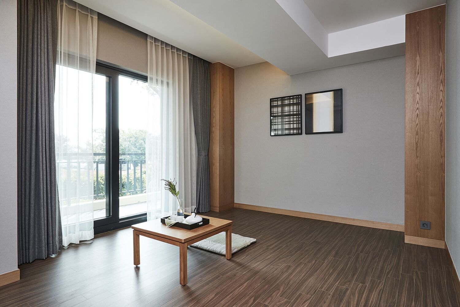 Una sala de estar moderna con piso de madera, un ventanal con cortinas ligeras, una mesita de madera con un florero, y dos obras de arte enmarcadas en la pared.