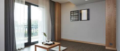 Una sala de estar moderna con piso de madera, un ventanal con cortinas ligeras, una mesita de madera con un florero, y dos obras de arte enmarcadas en la pared.