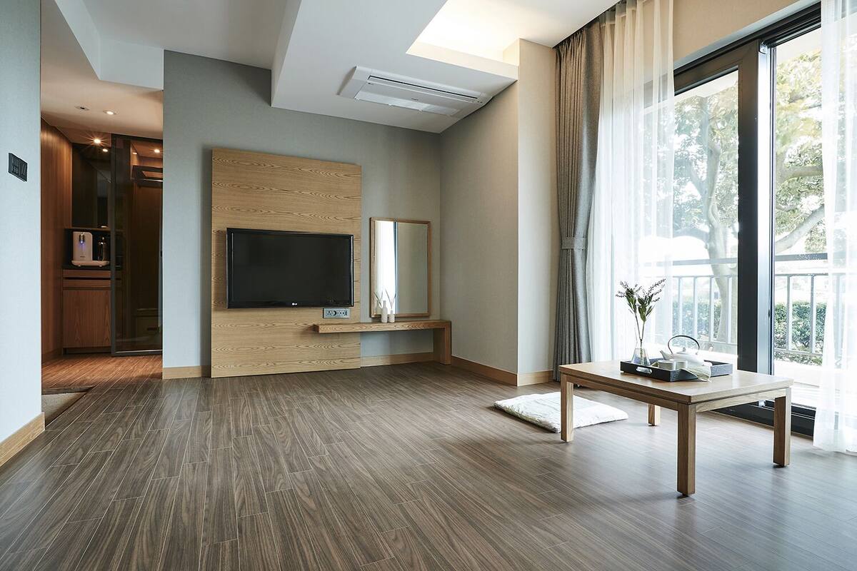 A modern living room with a flat-screen TV, wooden panel wall, large window with curtains, and a small table with a vase.
