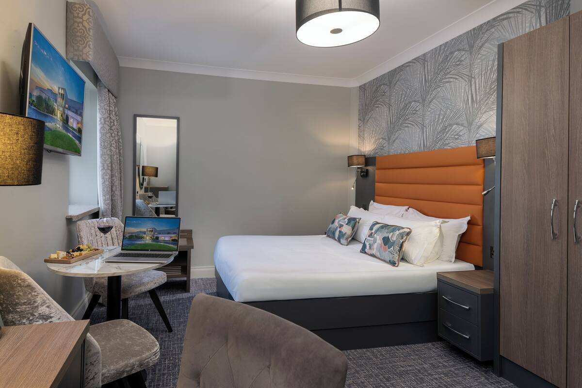 A hotel room with a bed, a desk with a laptop, a chair, a wardrobe, and a wall with a patterned design.