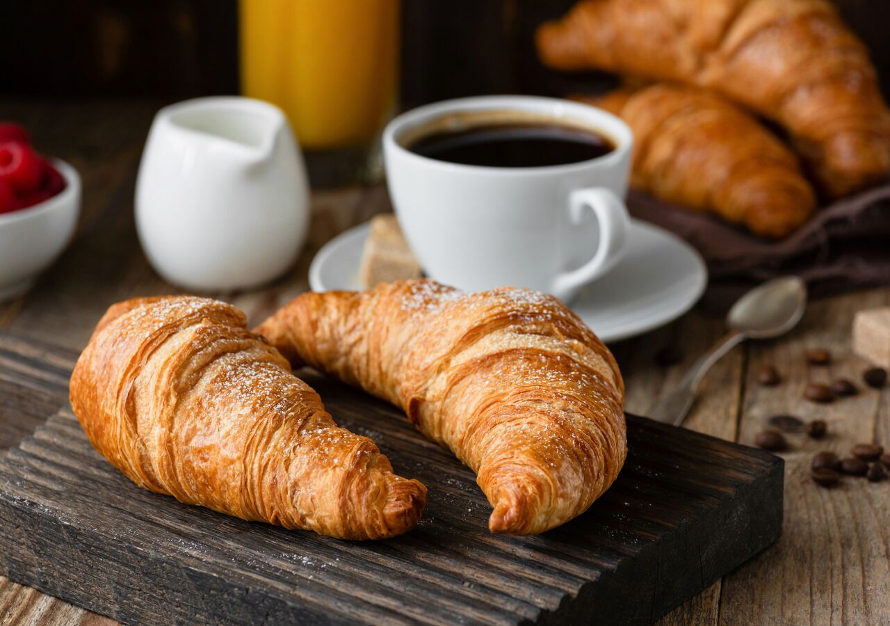 Dos croissants sobre una tabla de madera, una taza de café y un tazón pequeño de frambuesas.