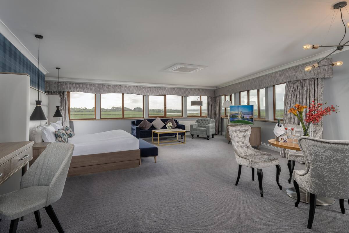 A hotel room with a bed, a desk, a sofa, a dining table with chairs, a TV, and large windows with a view.