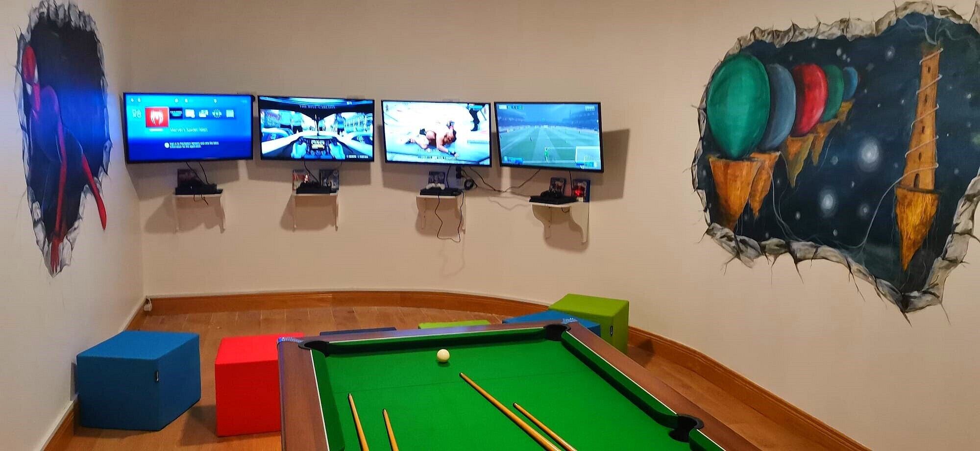 A room with a pool table, four monitors displaying different video games, and a wall mural.