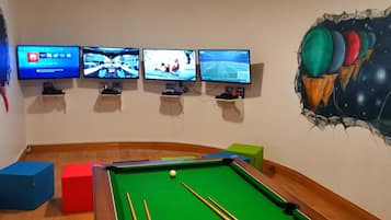 A room with a pool table, four monitors displaying different video games, and a wall mural.