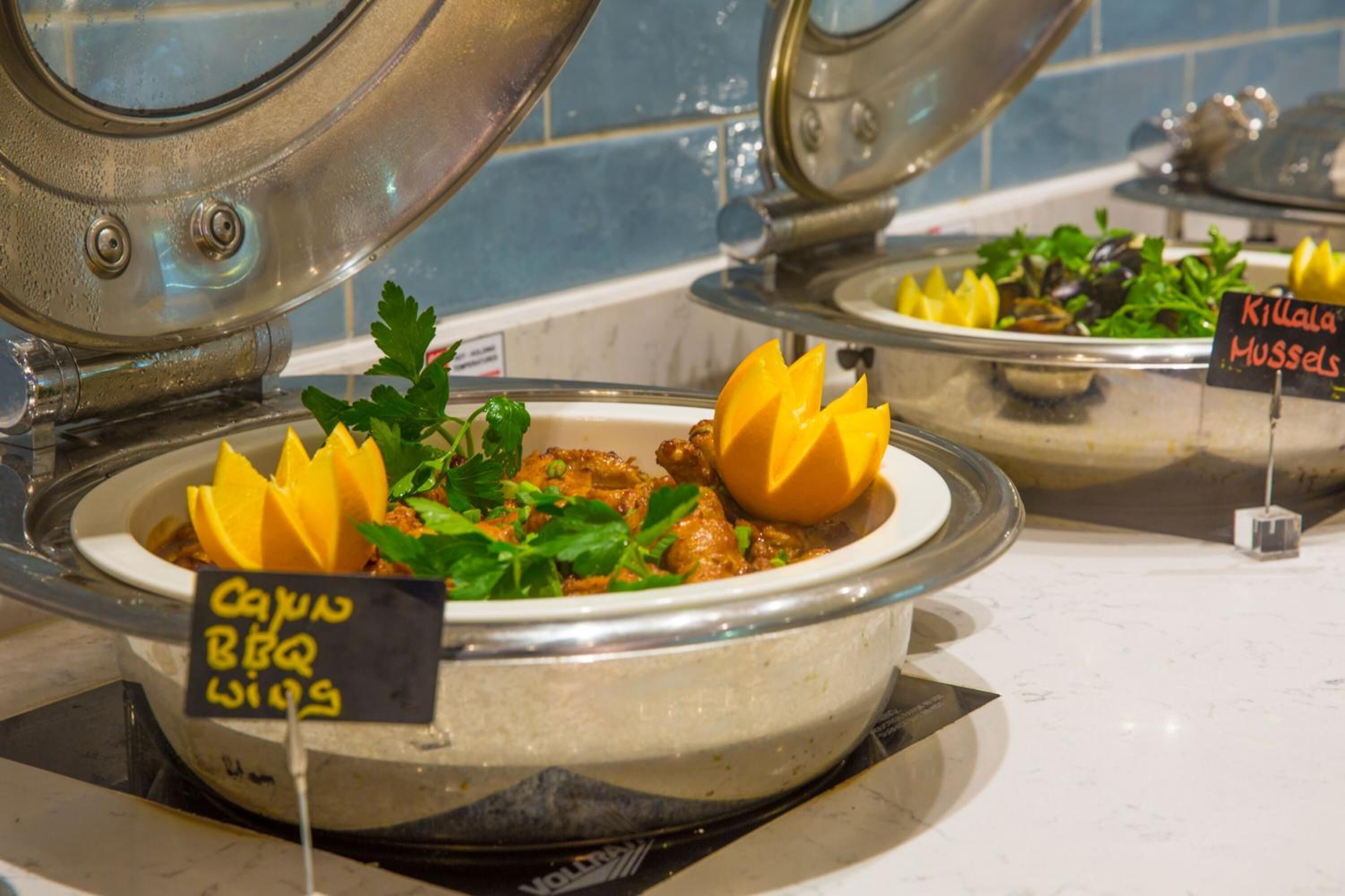 A buffet with labeled dishes including Cajun BBQ Wings and Kilaala Mussels.