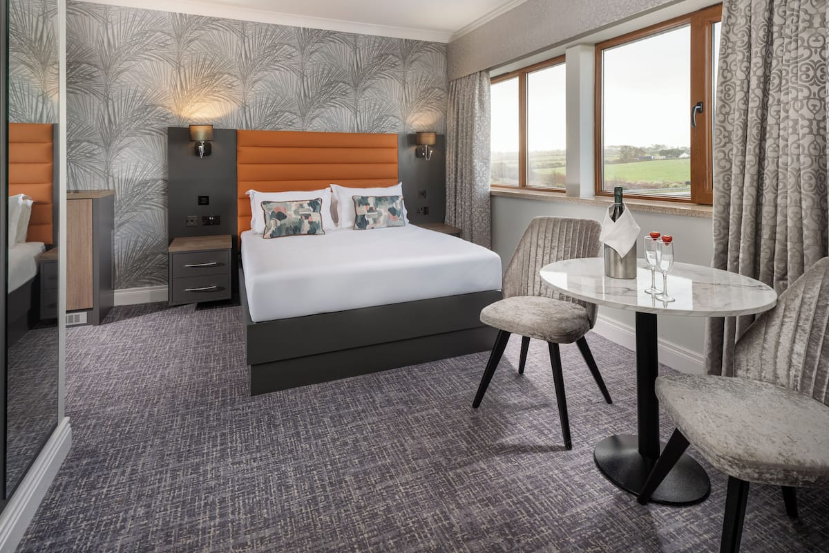 A hotel room with a bed, two bedside tables, a chair, a small round table with two glasses and a bottle, and a window with curtains.