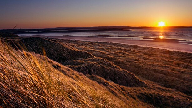 Un atardecer sobre una playa con dunas y un mar tranquilo.
