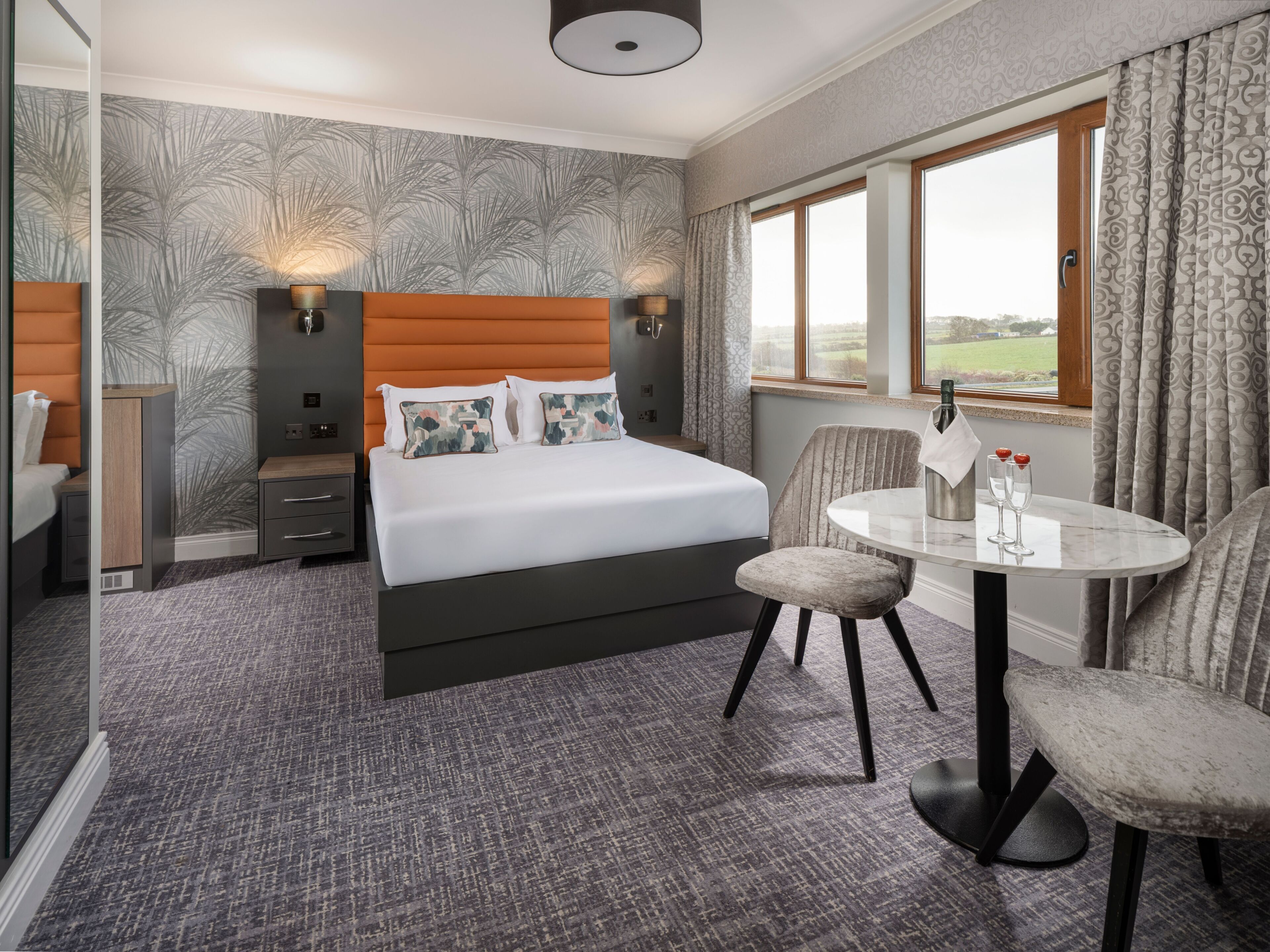 A hotel room with a bed, a small table with two chairs, a nightstand, a lamp, and a window with curtains.