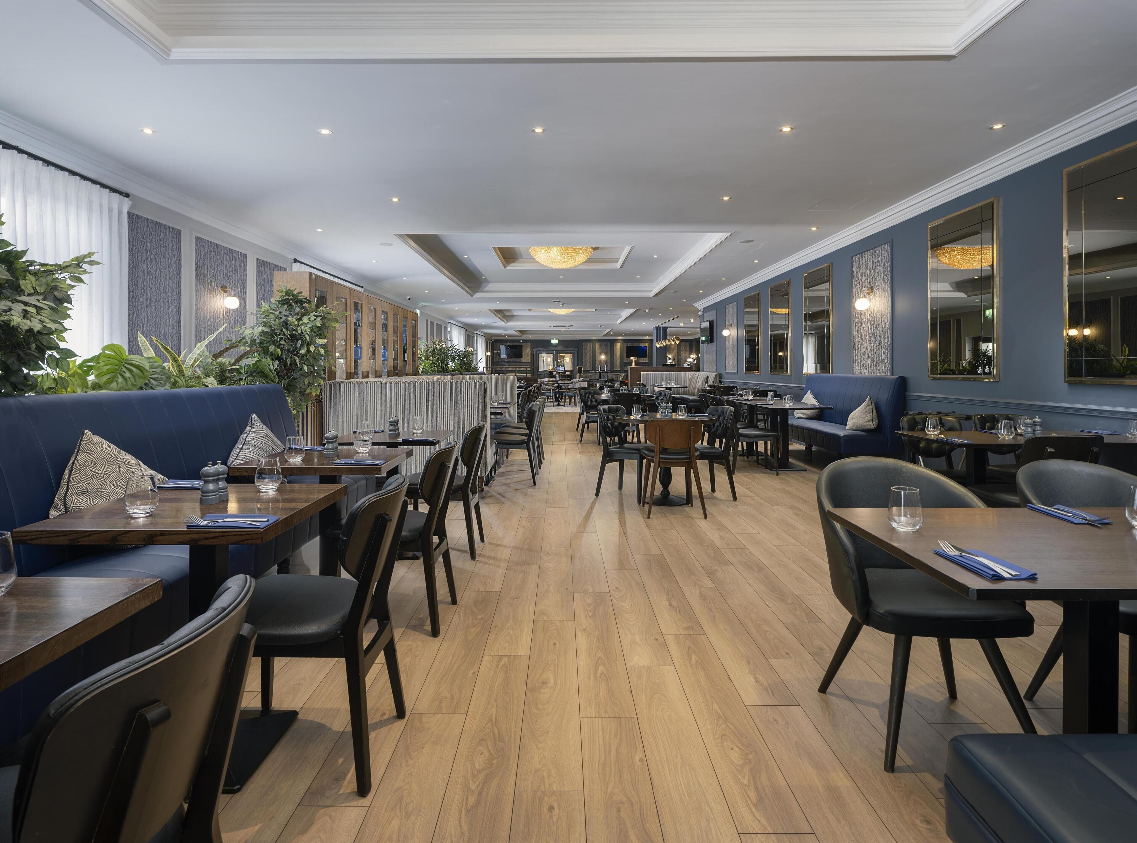 A spacious restaurant with wooden flooring, blue upholstered seating, and a variety of table settings.