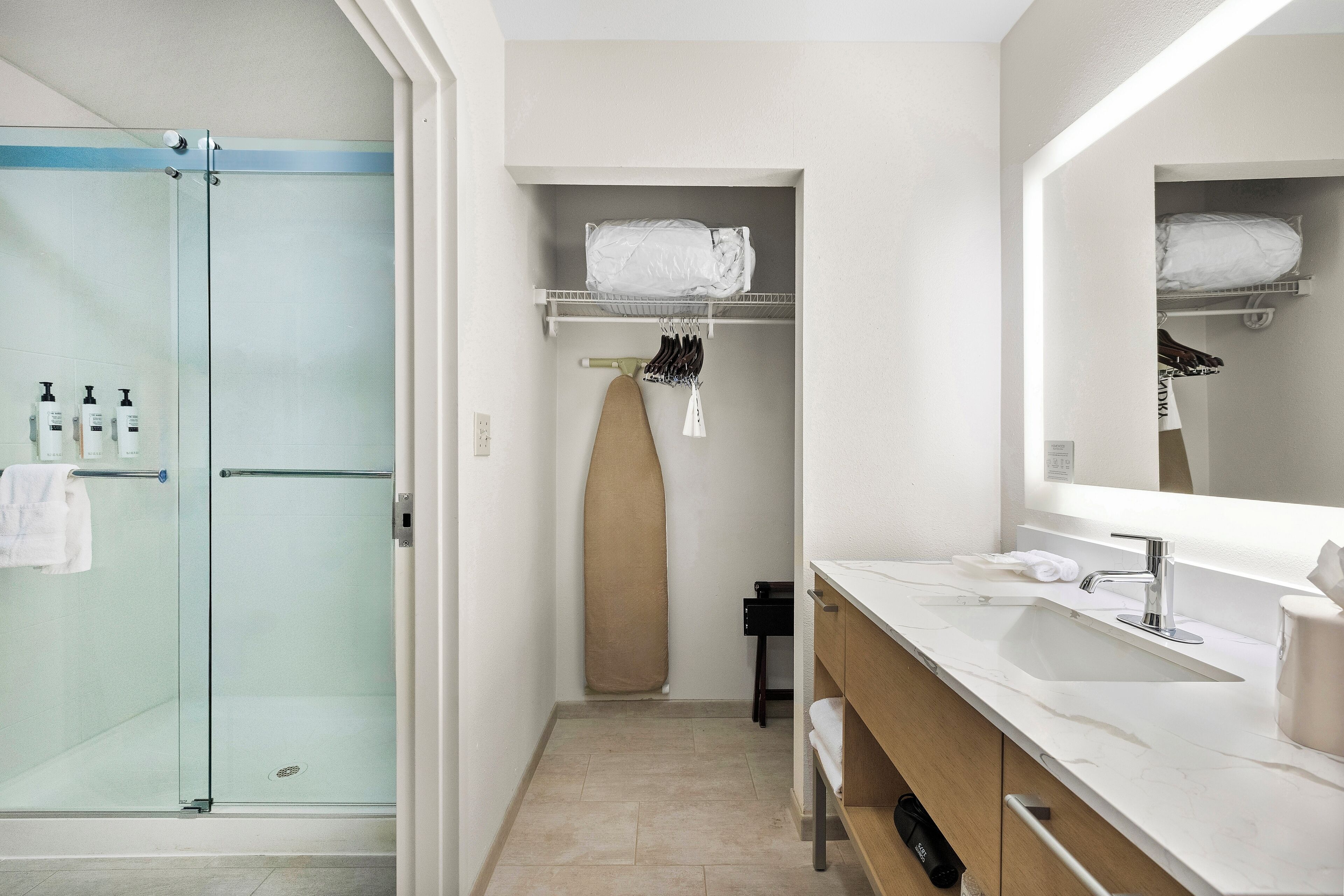 Une salle de bain moderne avec une douche à l’italienne en verre, un lavabo blanc avec un grand miroir et un placard de rangement avec des étagères.