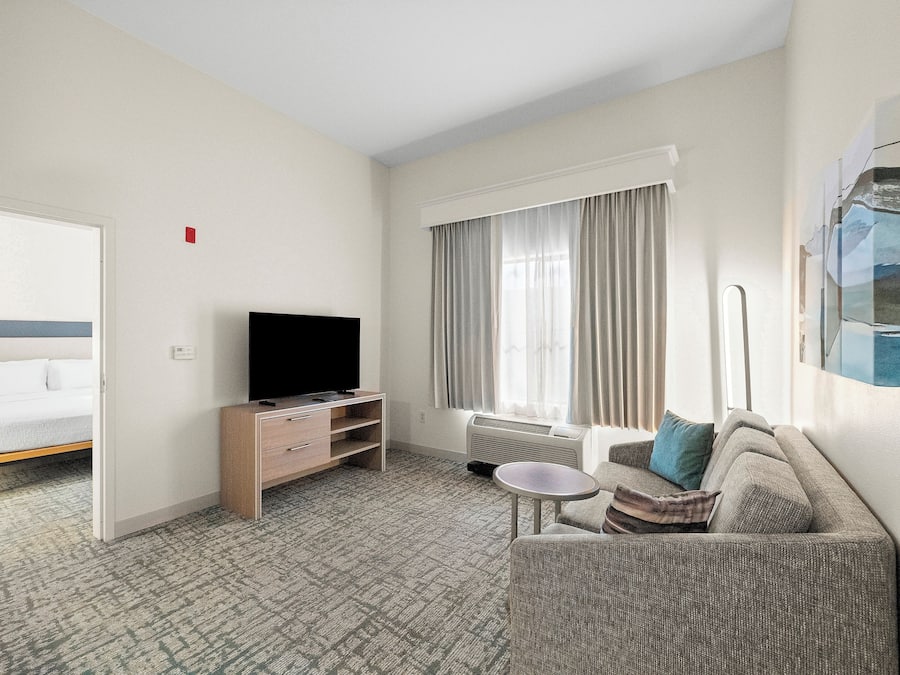 Habitación de hotel con cama, televisor sobre un soporte de madera, sofá, una mesita y una ventana con cortinas.