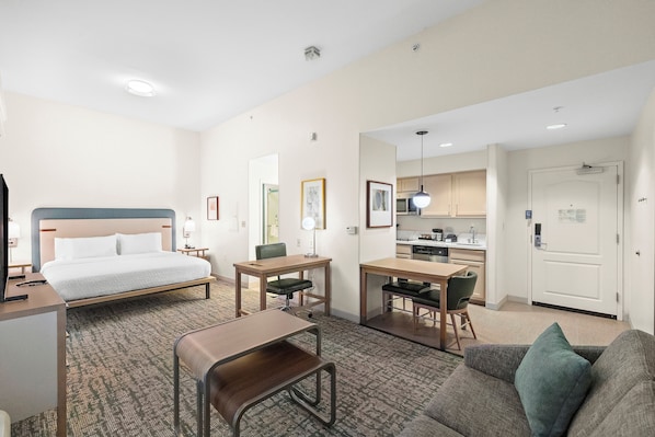 A hotel room with a bed, desk, chair, sofa, kitchenette, and a door.
