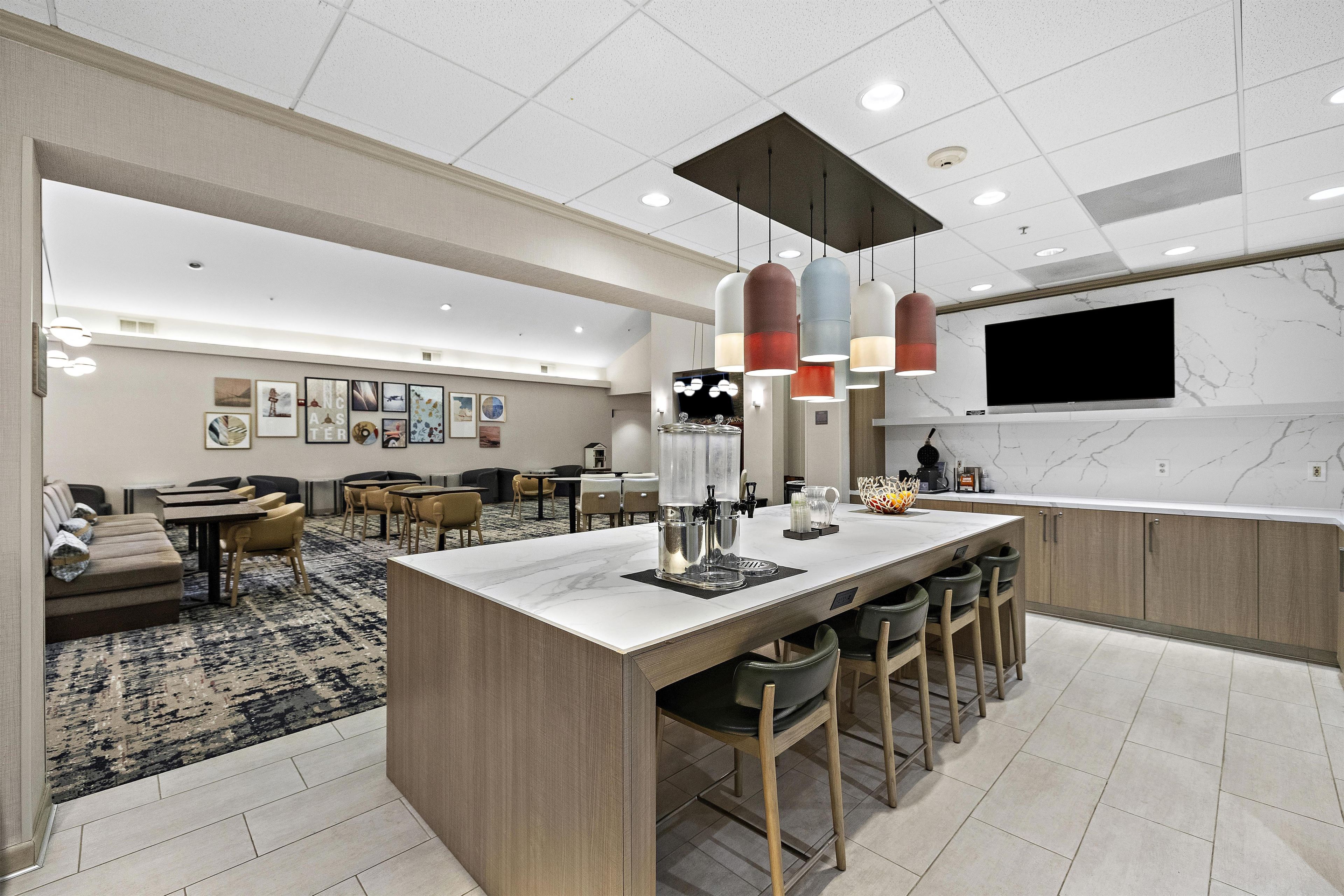 A modern hotel lobby with a central bar area, seating arrangements, and a large flat-screen TV mounted on the wall.