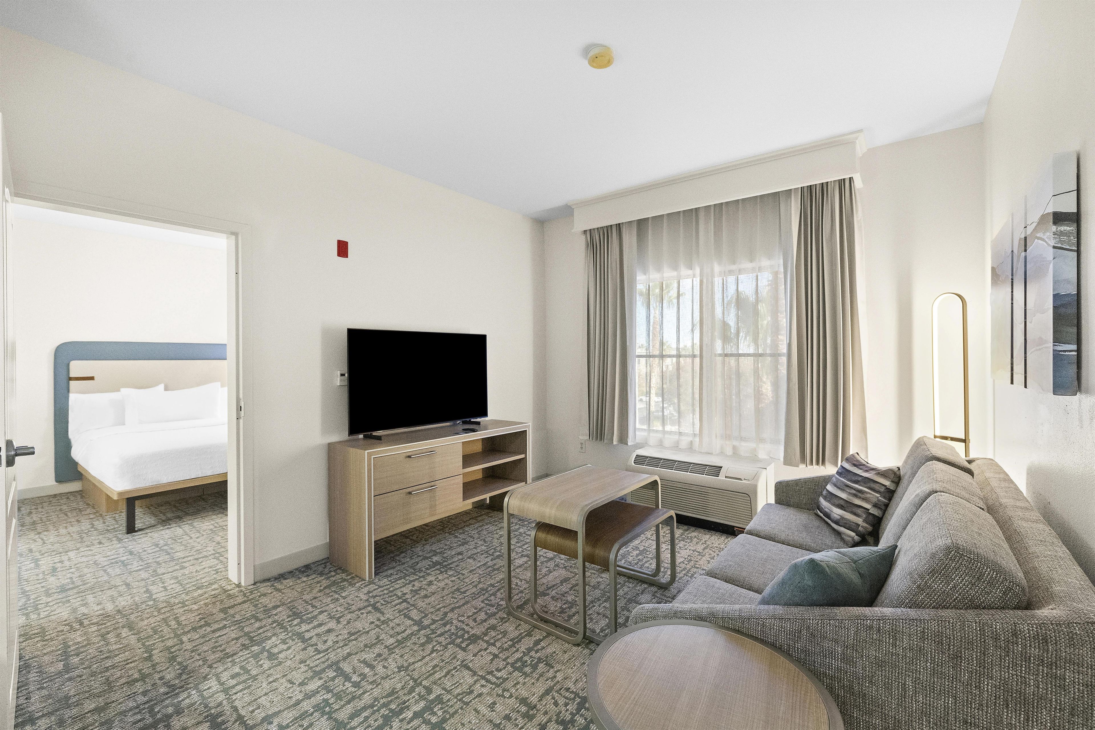 Quarto de hotel com cama, televisão, sofá, mesa pequena e janela com cortinas.