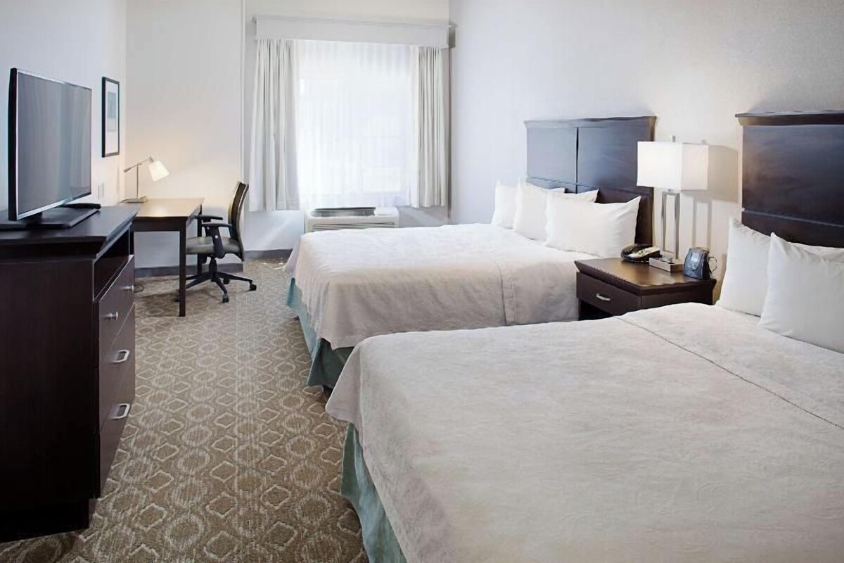 A hotel room with two beds, a desk with a chair, a television, and a window with curtains.