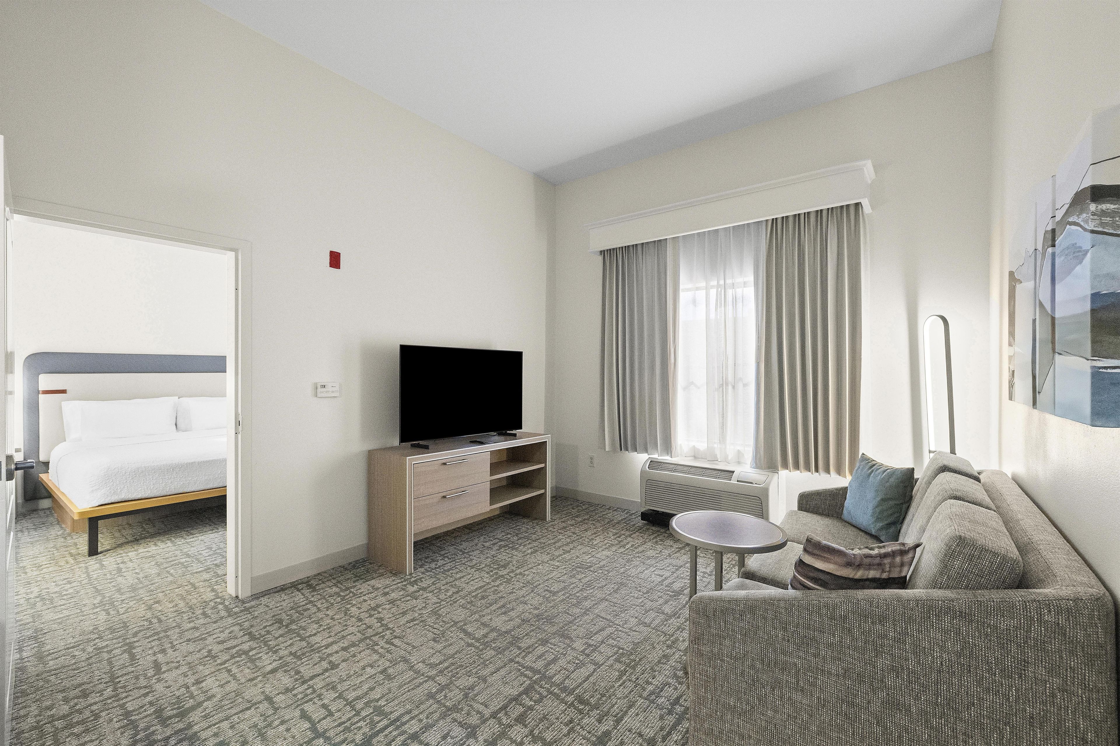 A hotel room with a bed, a TV on a stand, a sofa, a small table, and a window with curtains.