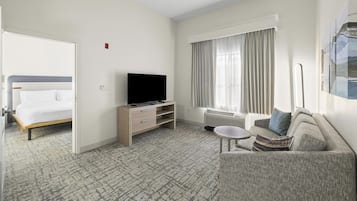 A hotel room with a bed, a TV on a stand, a sofa, a small table, and a window with curtains.