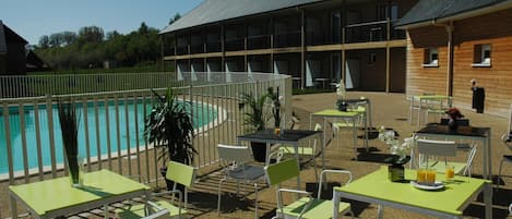 Uma área à beira da piscina com mesas e cadeiras, uma piscina e um edifício com várias varandas.