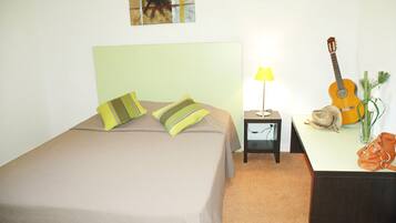 A neatly arranged hotel room with a bed, a bedside lamp, a guitar, and a small table with a vase and a handbag.