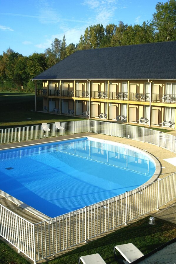 Una piscina con valla blanca, rodeada de sillas de descanso y un edificio con balcones.