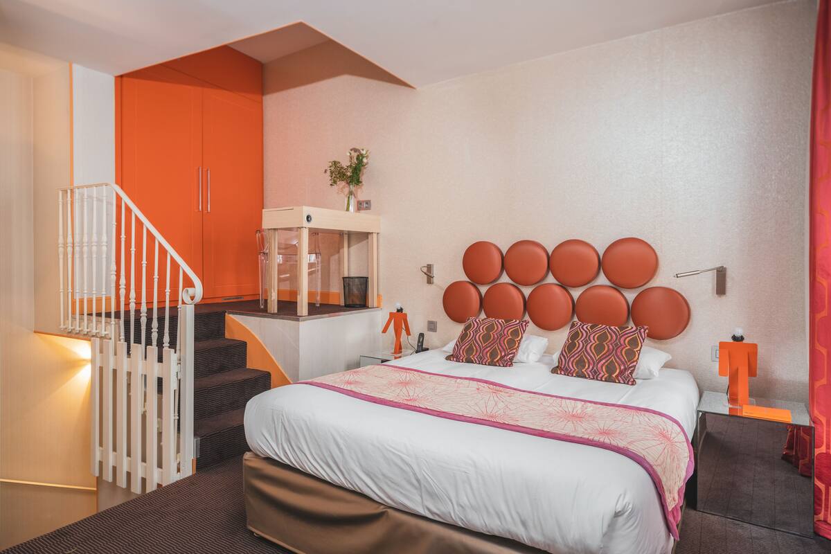 A modern hotel room with a large bed, a staircase, a small balcony, and a bathroom with a mirror.