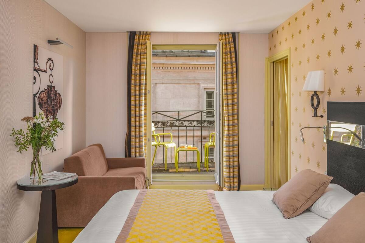 A hotel room with a bed, a sitting area with a chair, a small table with a vase, and a view of an outdoor balcony with yellow chairs.