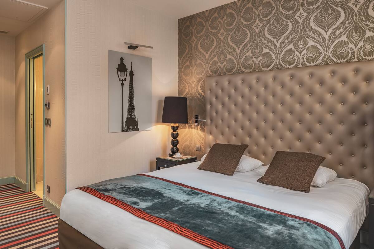 A hotel room with a large bed, a tufted headboard, a patterned wallpaper, a framed picture of the Eiffel Tower, and a striped carpet.