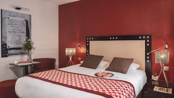 Habitación de hotel con una cama grande, una mesita con un florero y un libro, una silla y una mesita de noche con una lámpara y un teléfono.