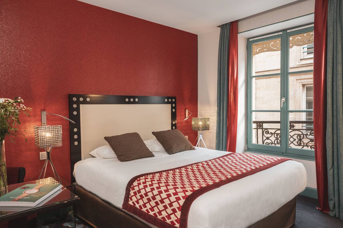 A hotel room with a large bed, red walls, a window with curtains, and a small table with a lamp and a book.