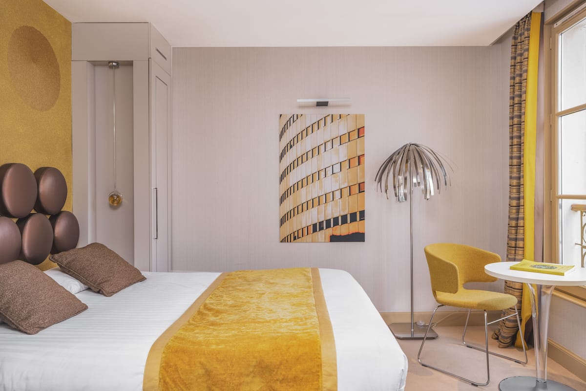 A hotel room with a large bed, a small round table, a chair, a floor lamp, and a painting on the wall.