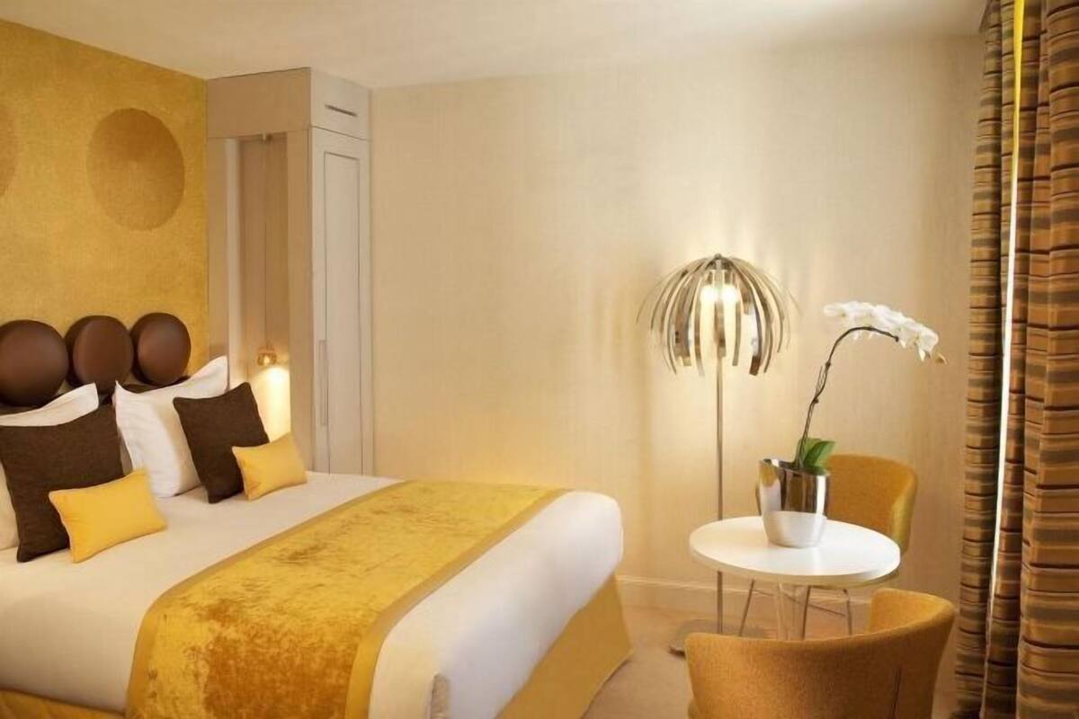 A hotel room with a bed, a bedside table with a lamp, a chair, and a potted plant.
