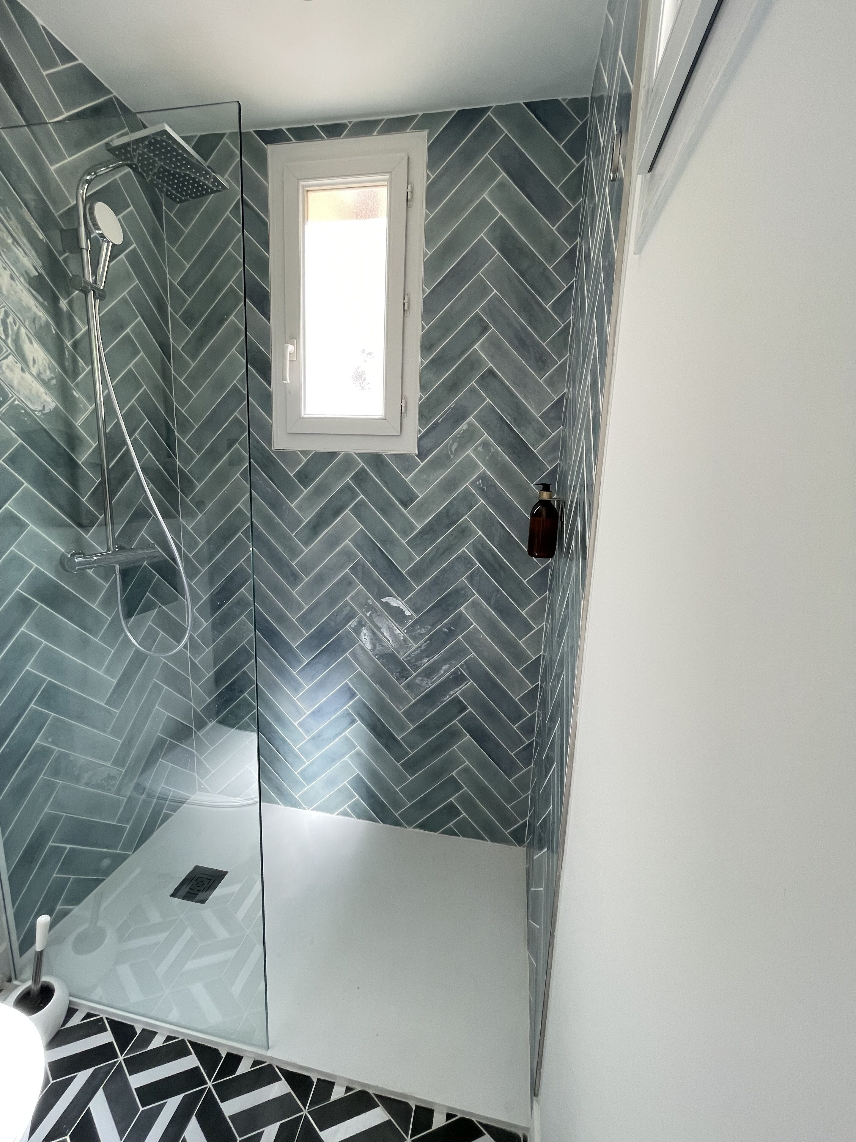 Une douche moderne avec une porte en verre, une petite fenêtre et un motif de carrelage en chevrons sur les murs.