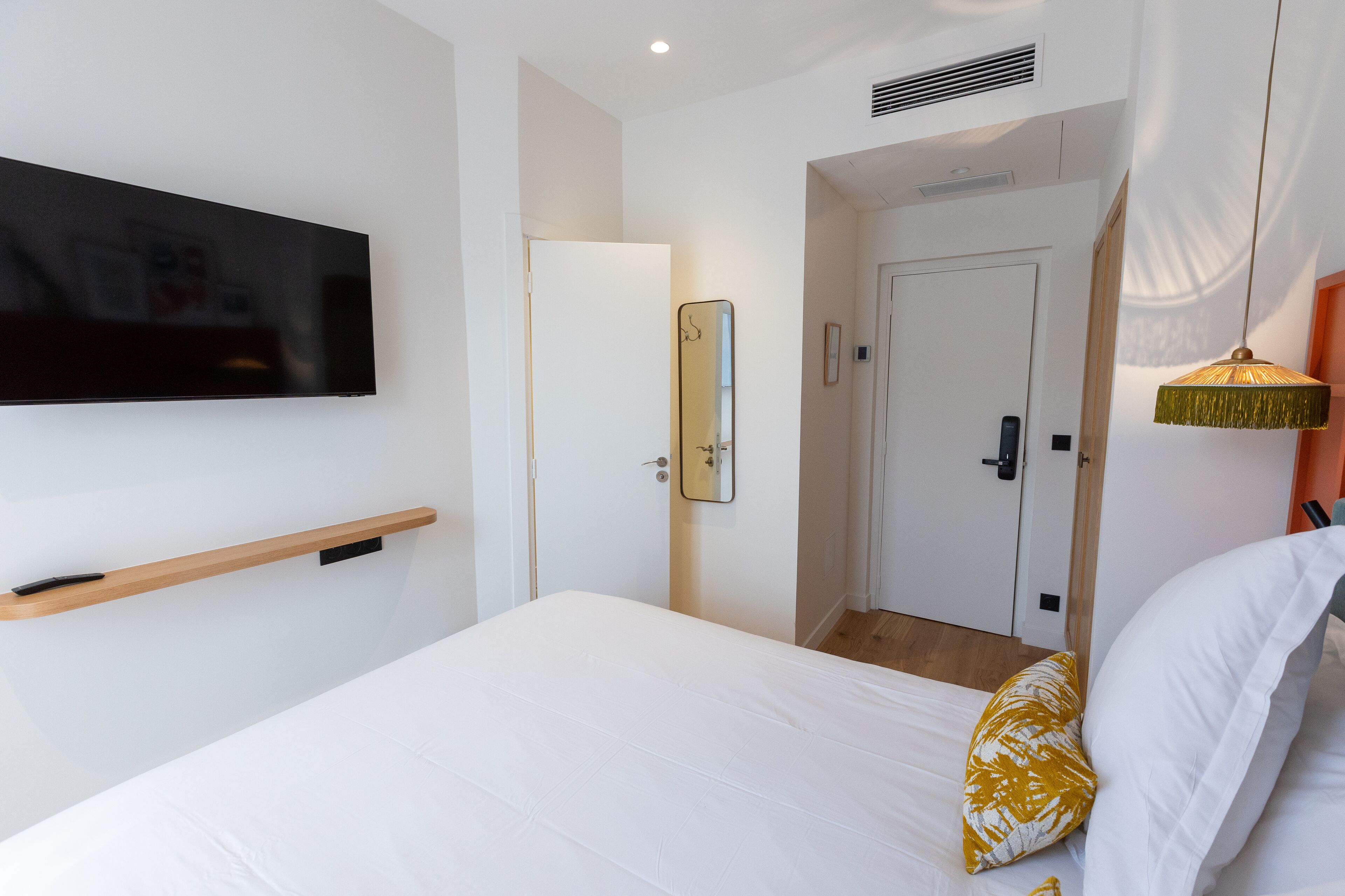 Una habitación de hotel con una cama, un televisor montado, una puerta y un pasillo.