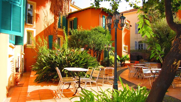 A courtyard with outdoor seating, a street lamp, and a building with orange walls and green shutters. - Maison Durante (Nice)