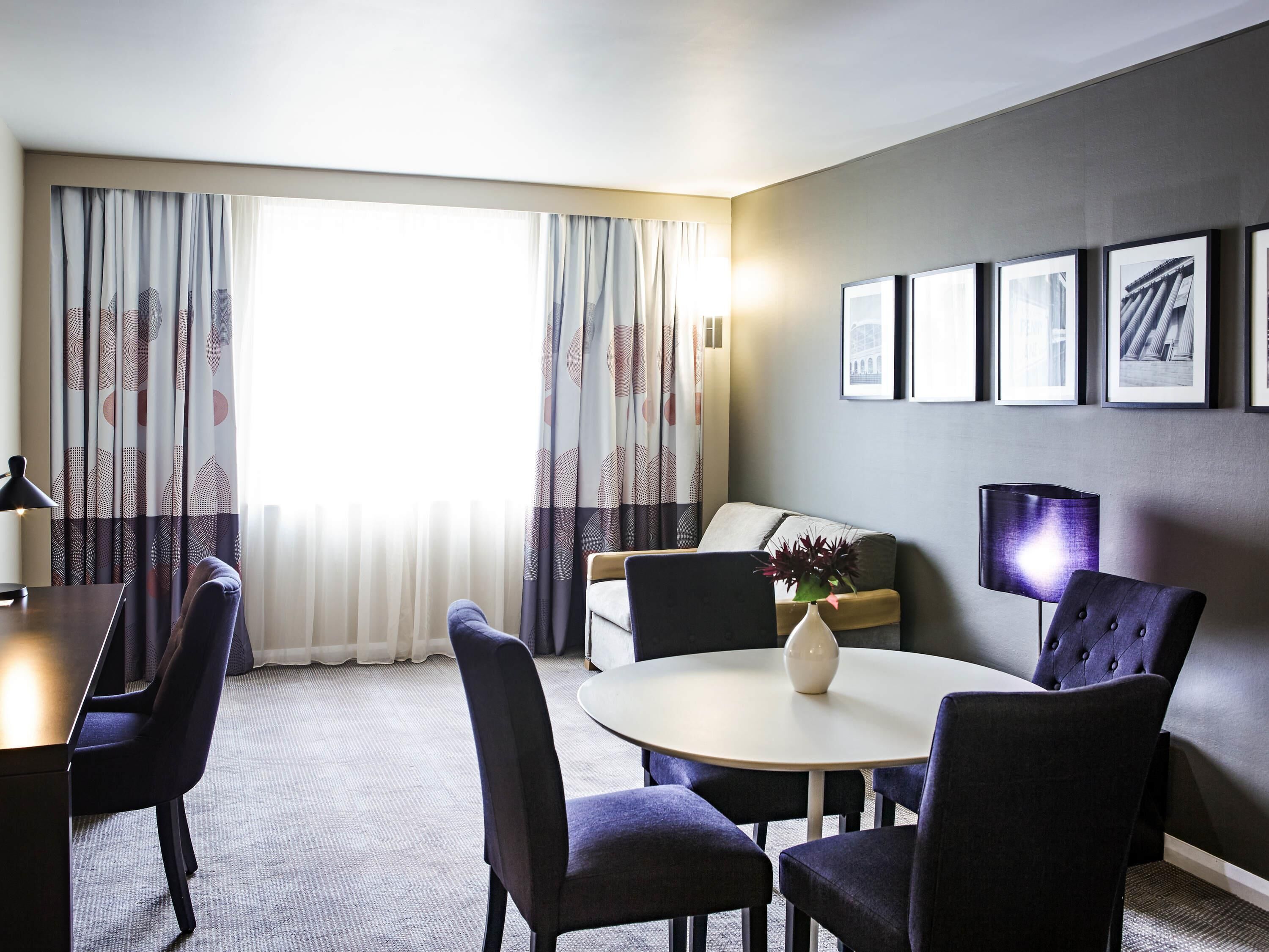 A hotel room with a dining table, chairs, a sofa, a lamp, and a vase with flowers.