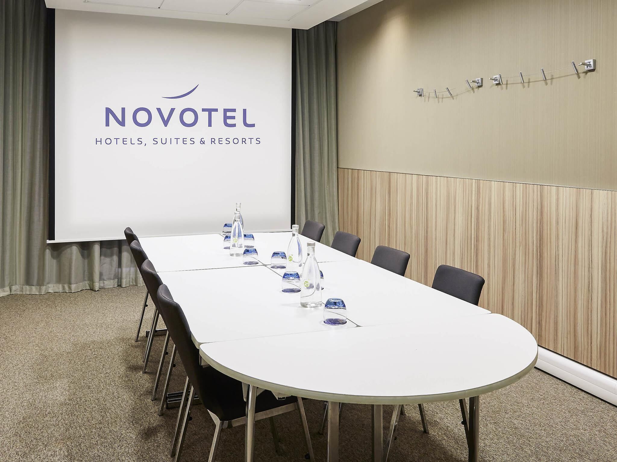 A conference room with a long table, chairs, a projector screen displaying 'NOVOTEL HOTELS, SUITES & RESORTS', and a wooden panel wall.