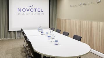 A conference room with a long table, chairs, a projector screen displaying 'NOVOTEL HOTELS, SUITES & RESORTS', and a wooden panel wall.