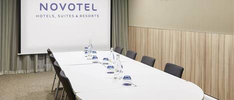 A conference room with a long table, chairs, a projector screen displaying 'NOVOTEL HOTELS, SUITES & RESORTS', and a wooden panel wall.