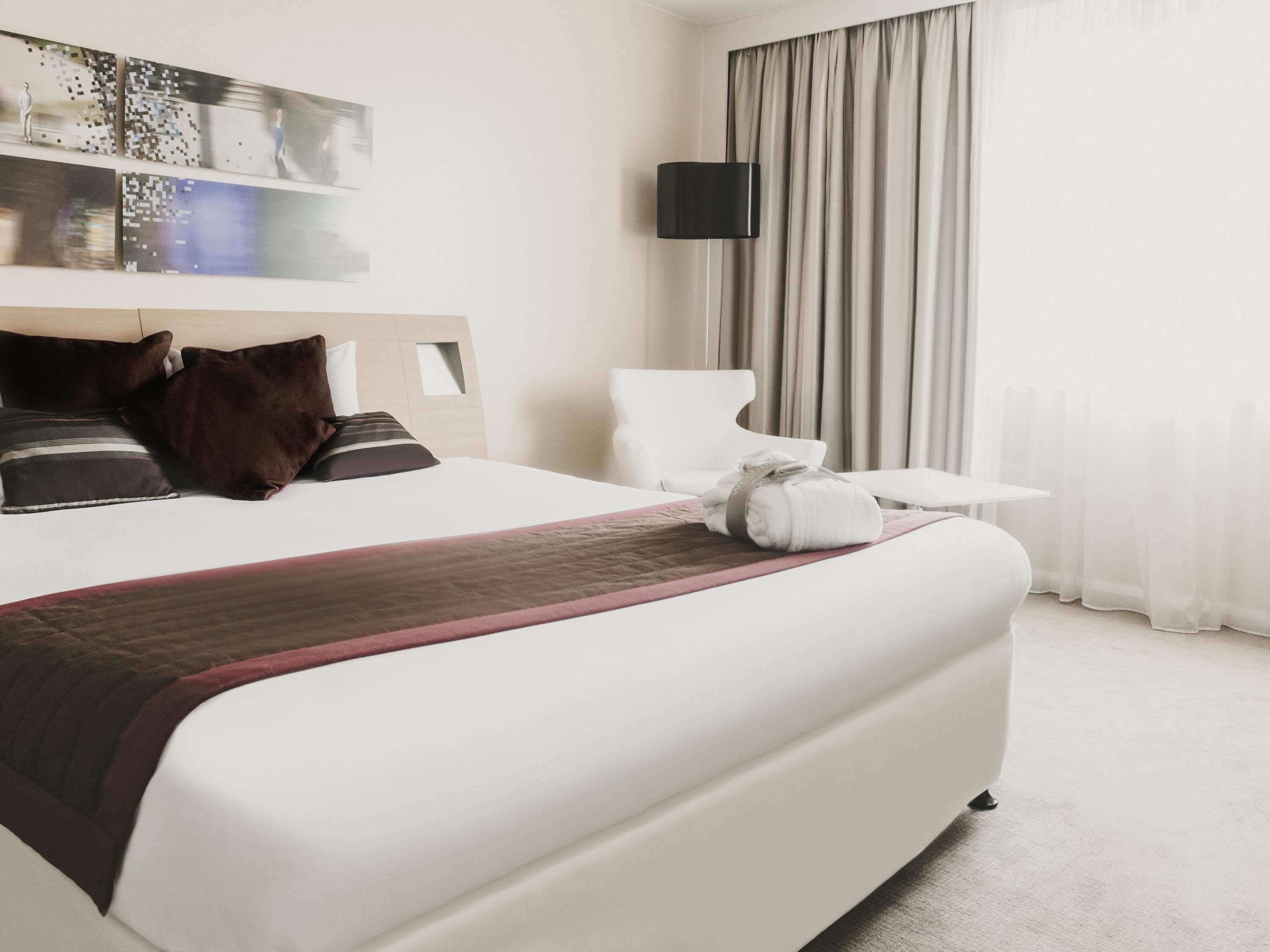A hotel room with a large bed, a flat-screen TV mounted on the wall, a chair, and a window with curtains.