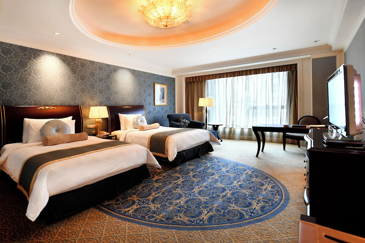 A hotel room with two beds, a desk, a chair, a television, and a large window with curtains.