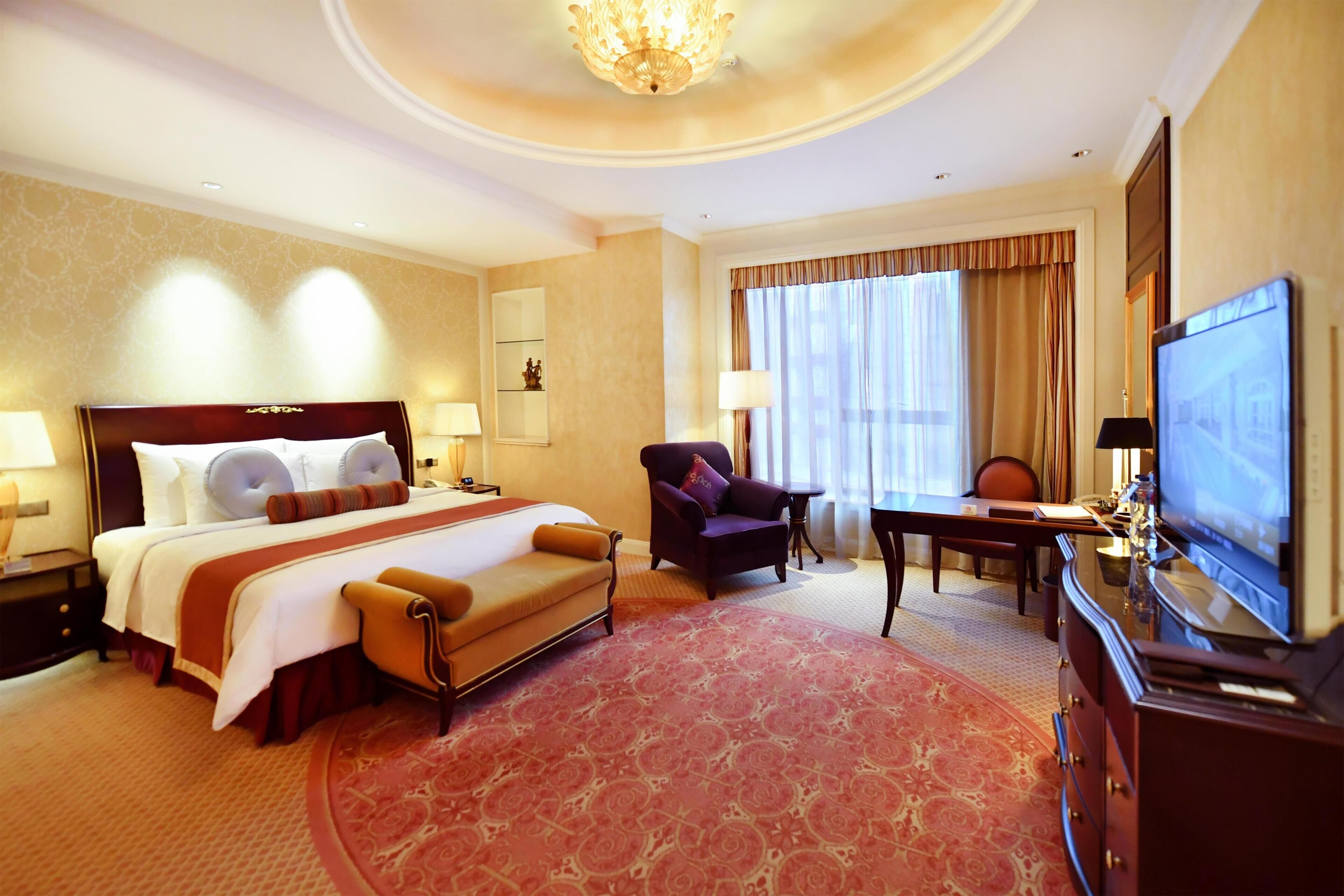 A hotel room with a large bed, a desk, a chair, a television, and a chandelier.
