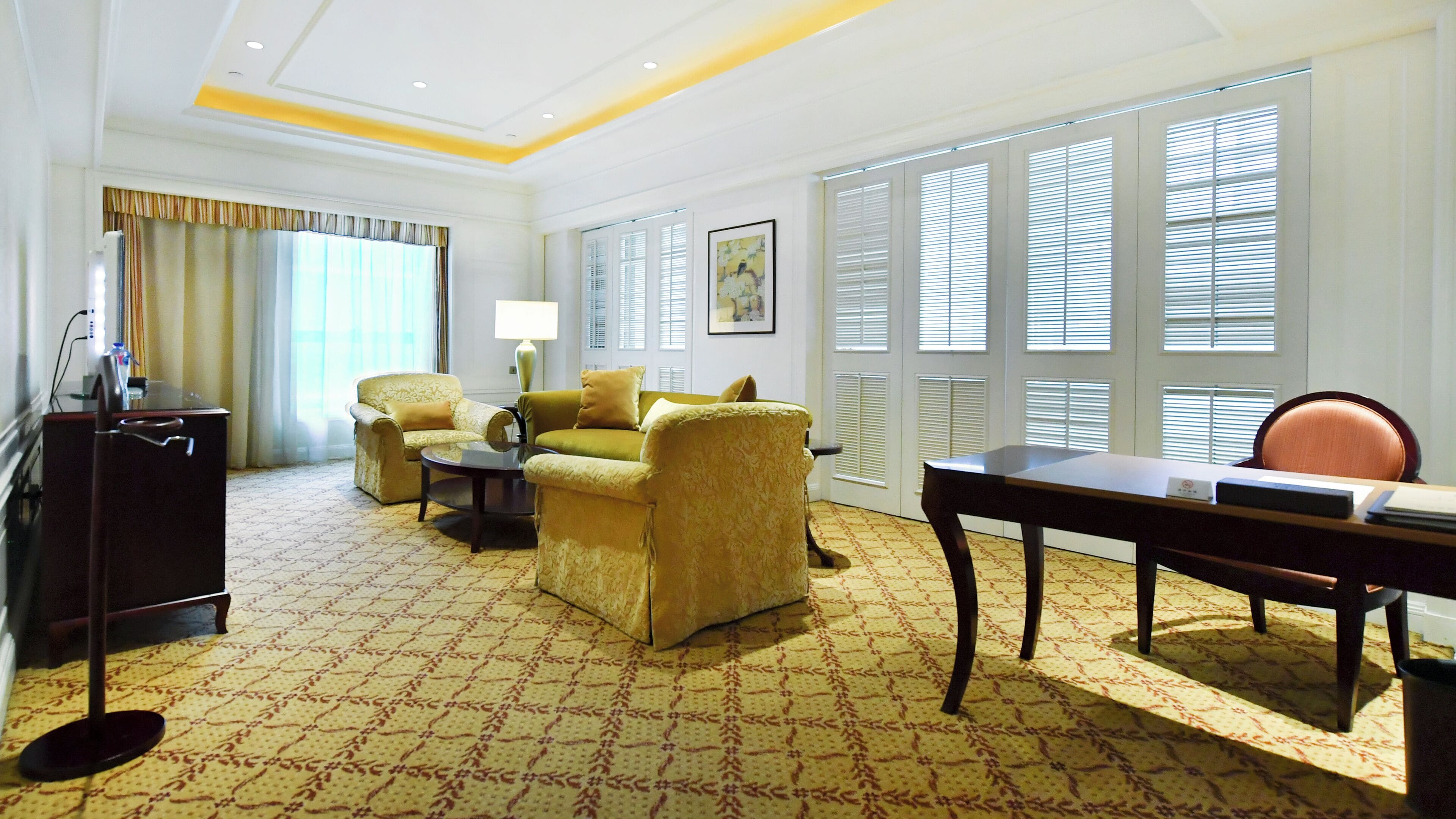 A hotel room with a patterned carpet, a sofa, a chair, a desk, and a lamp.