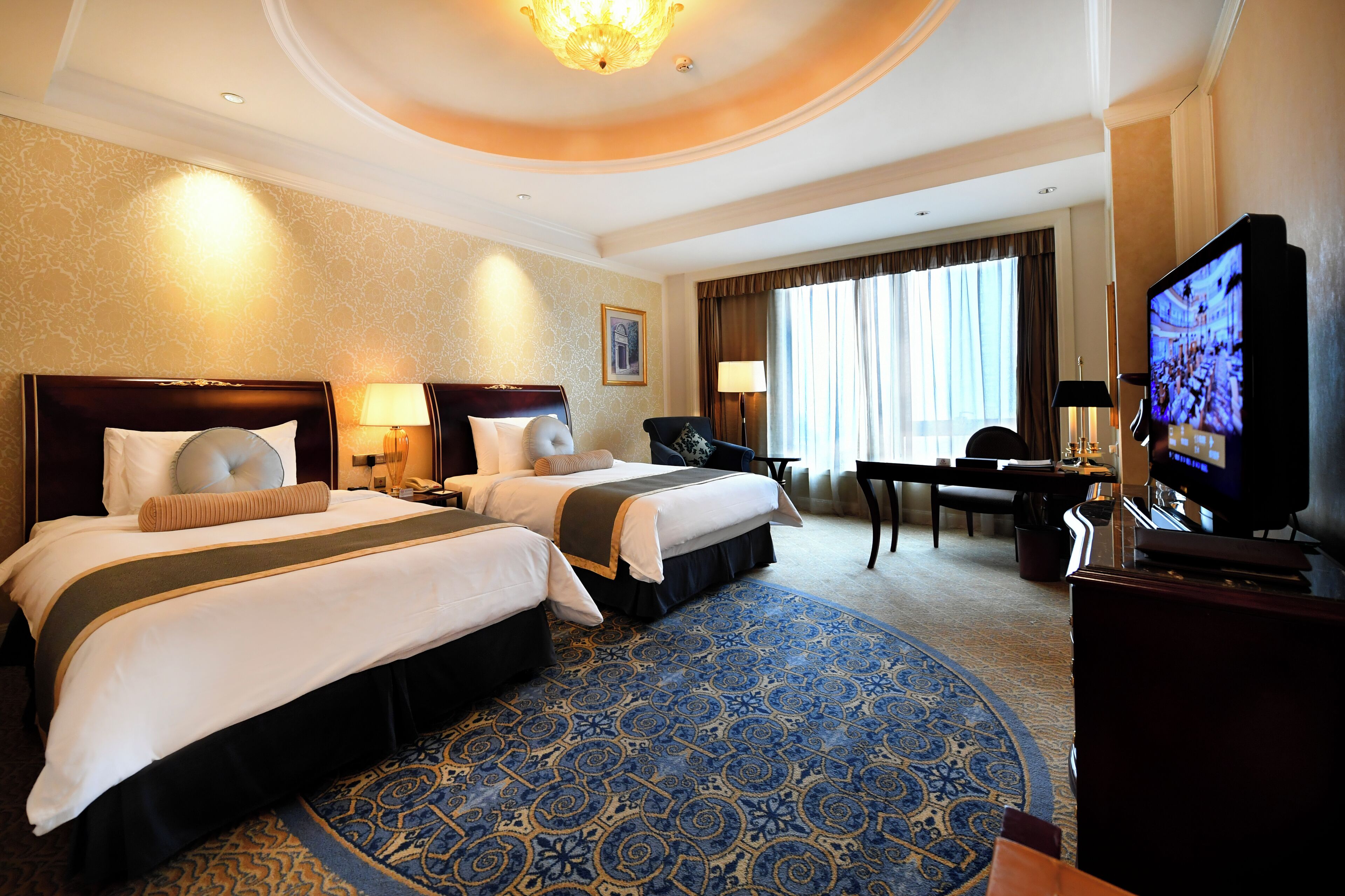 A hotel room with two beds, a television, a desk, and a large window with curtains.