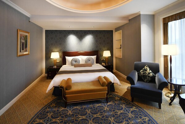 Habitación de hotel con una cama grande, zona de estar con una silla y mesa, alfombra estampada y un cuadro en la pared.