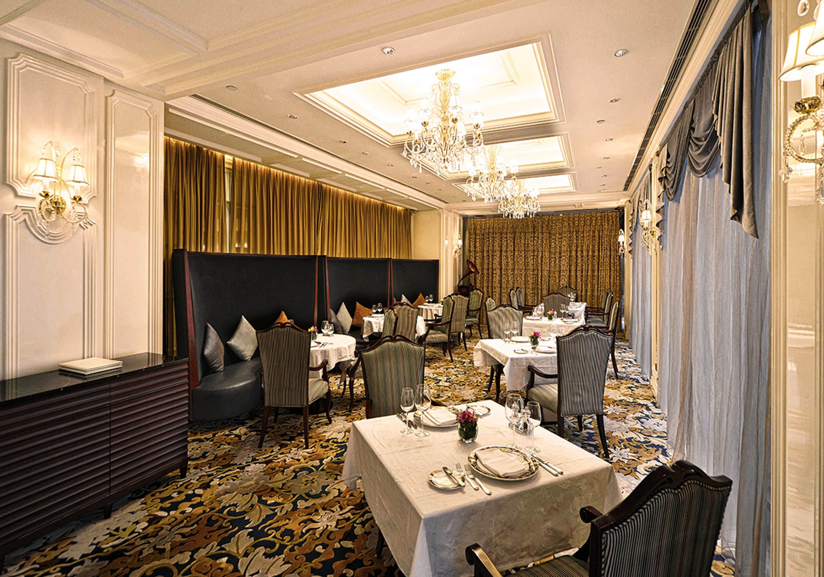 A formal dining room with tables set for service, elegant chandeliers, and a patterned carpet.