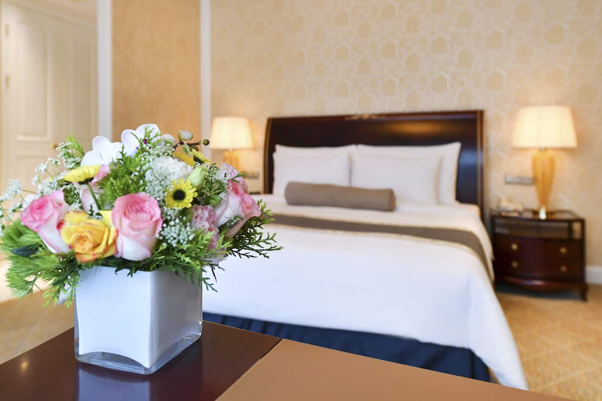 A hotel room with a neatly made bed, a bedside table with a vase of flowers, and a nightstand with a lamp.
